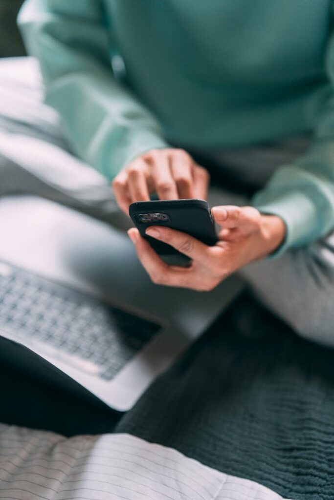 Crop anonymous male freelancer text messaging on modern cellphone while sitting on bed with laptop during remote work at home